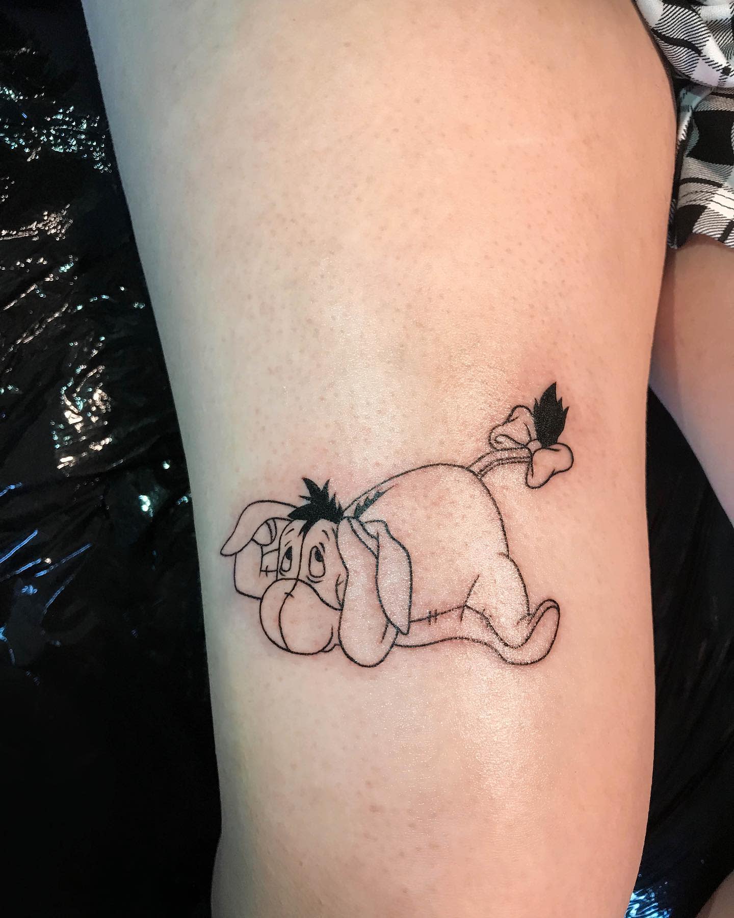Tattoo of Eeyore from Winnie the Pooh, lying down with a sad expression on a person's thigh